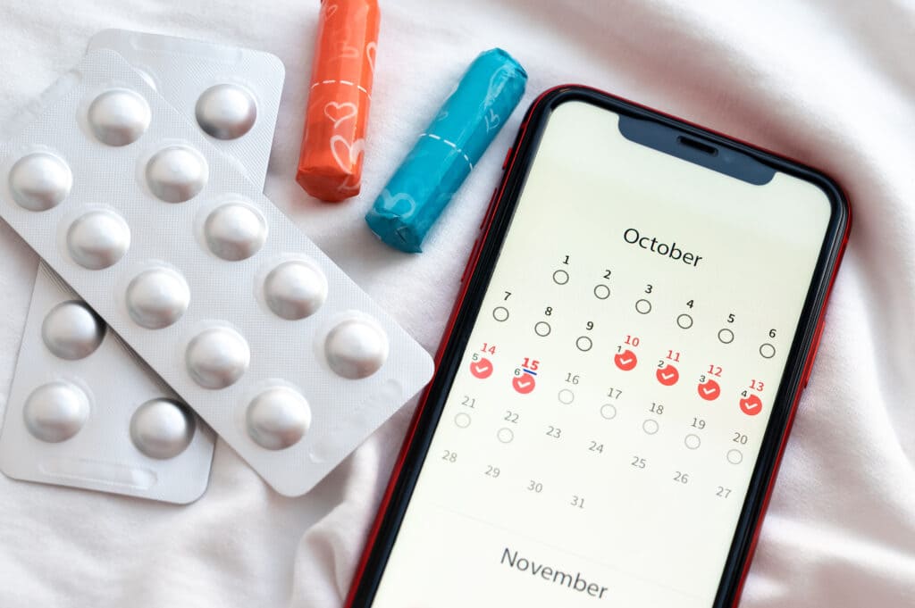 A smartphone displays a period tracking calendar for October, ideal for scripture-based cycle syncing. Nearby are blister packs of pills and three tampons, all placed on a white fabric surface.