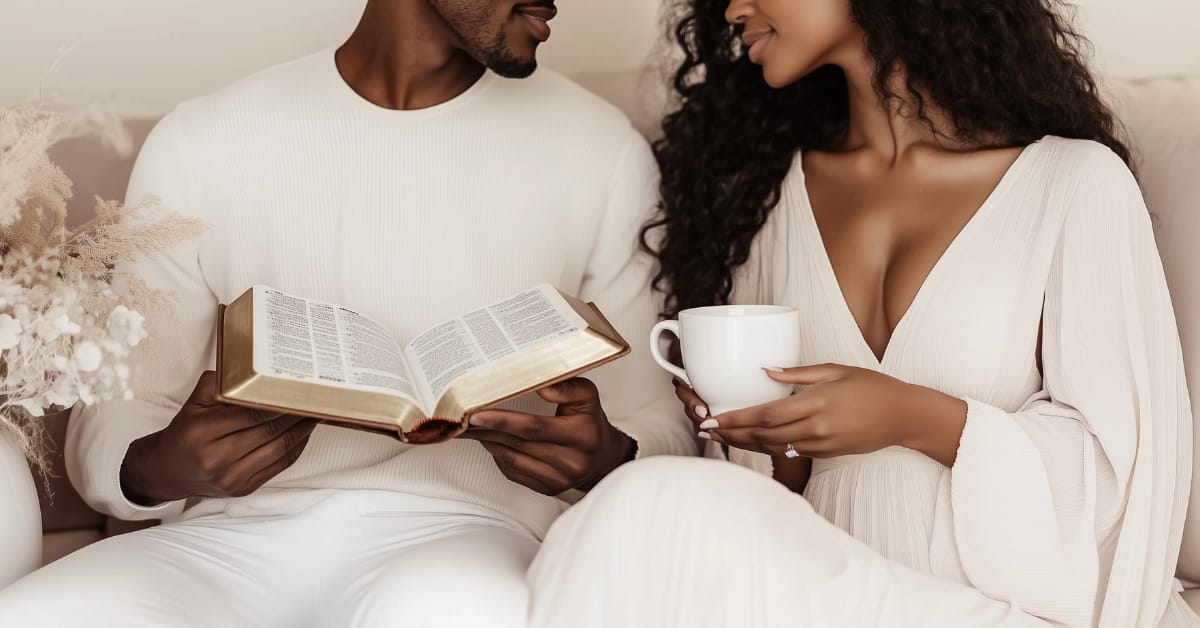 A couple sits closely together on a couch. The man holds an open book of Biblical Wisdom for Building Strong Lasting Relationships, while the woman holds a white mug. They smile at each other, wearing matching light-colored, comfortable clothing.