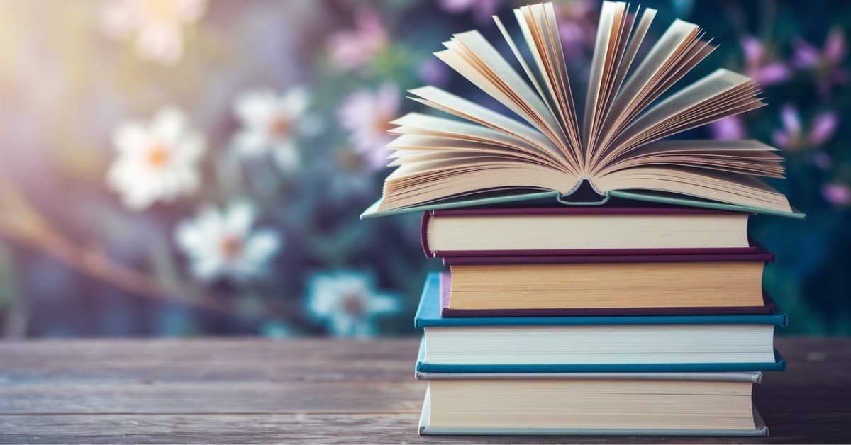 A stack of colorful books on a wooden table with one book open on top. The background features blurred pink and white flowers. The text becomingandbloom.com with a leaf logo is visible in the lower-left corner.
