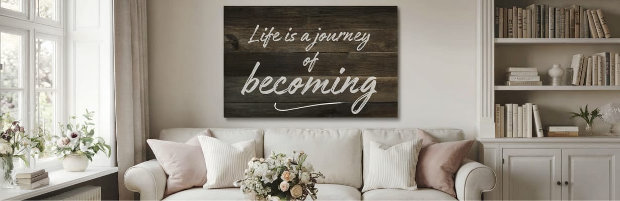 A cozy living room with a white couch, pastel cushions, floral arrangements, shelves with books and decor, and a wooden wall sign that reads, “Life is a journey of becoming.”.