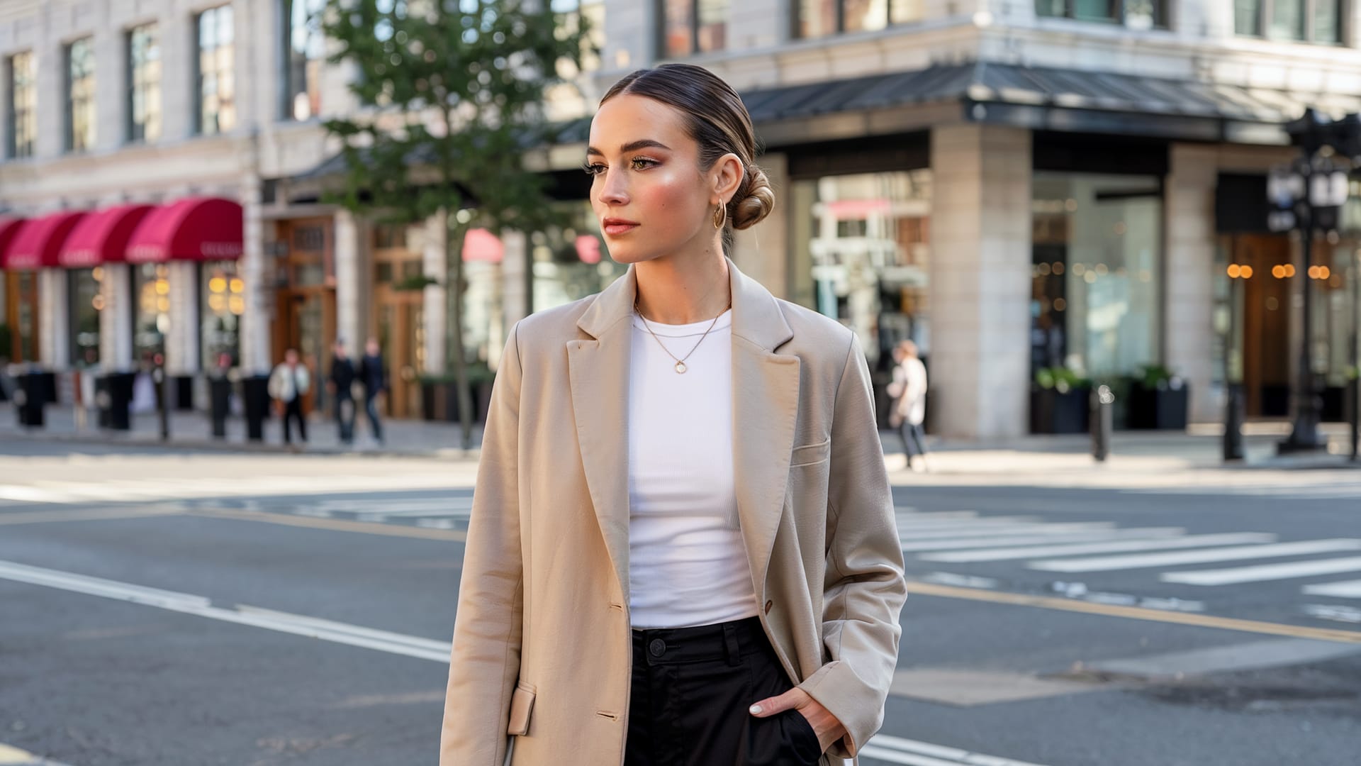 A woman wearing a beige blazer and white shirt stands on a city street corner, embodying the timeless style seen throughout the history of Clean Girl Aesthetic. Shops and pedestrians are visible in the background on a sunny day.