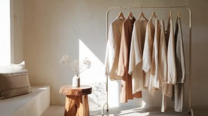 A serene, natural-light filled dressing area with a curated rack of sustainable luxury garments in neutral tones—cream linens, camel wools, and textured cottons. A handcrafted wooden stool holds a small vase with delicate dried flowers. The scene embodies mindful consumption with quality over quantity.
