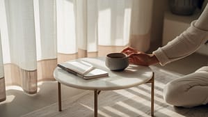 A person in light clothing sits by a small round table with a notebook and pen, reaching for a ceramic mug. Sunlight streams through sheer curtains, creating a calm, cozy atmosphere.