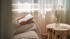 A person sitting peacefully by a window with a journal in their lap, soft morning light streaming in through sheer curtains. The scene has a calm, reflective quality with neutral tones of cream and soft blue. A small vase with delicate wildflowers sits nearby on a natural wood side table, symbolizing growth and beauty emerging from simplicity.