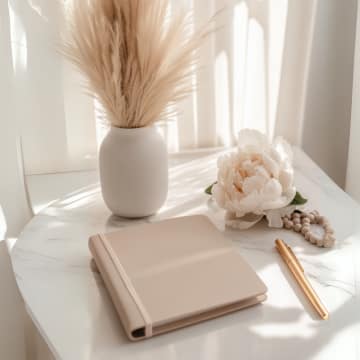 A beige notebook and a gold pen rest on a white marble table beside a white vase with pampas grass and a blooming white peony, all bathed in soft natural light.